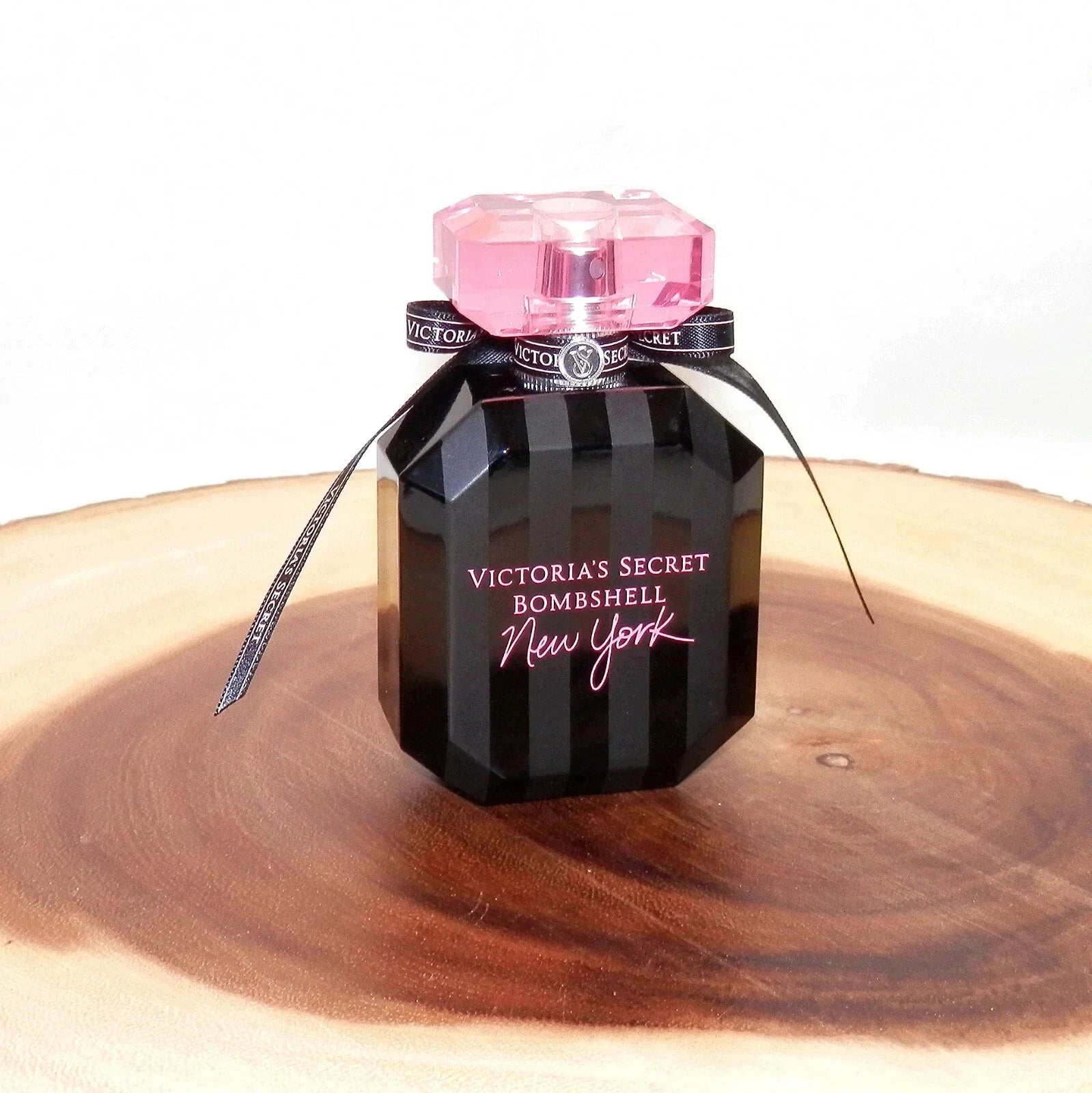 A black, faceted bottle of Victoria’s Secret Bombshell New York perfume with a pink crystal-style cap and ribbon accents, displayed on a wooden surface.